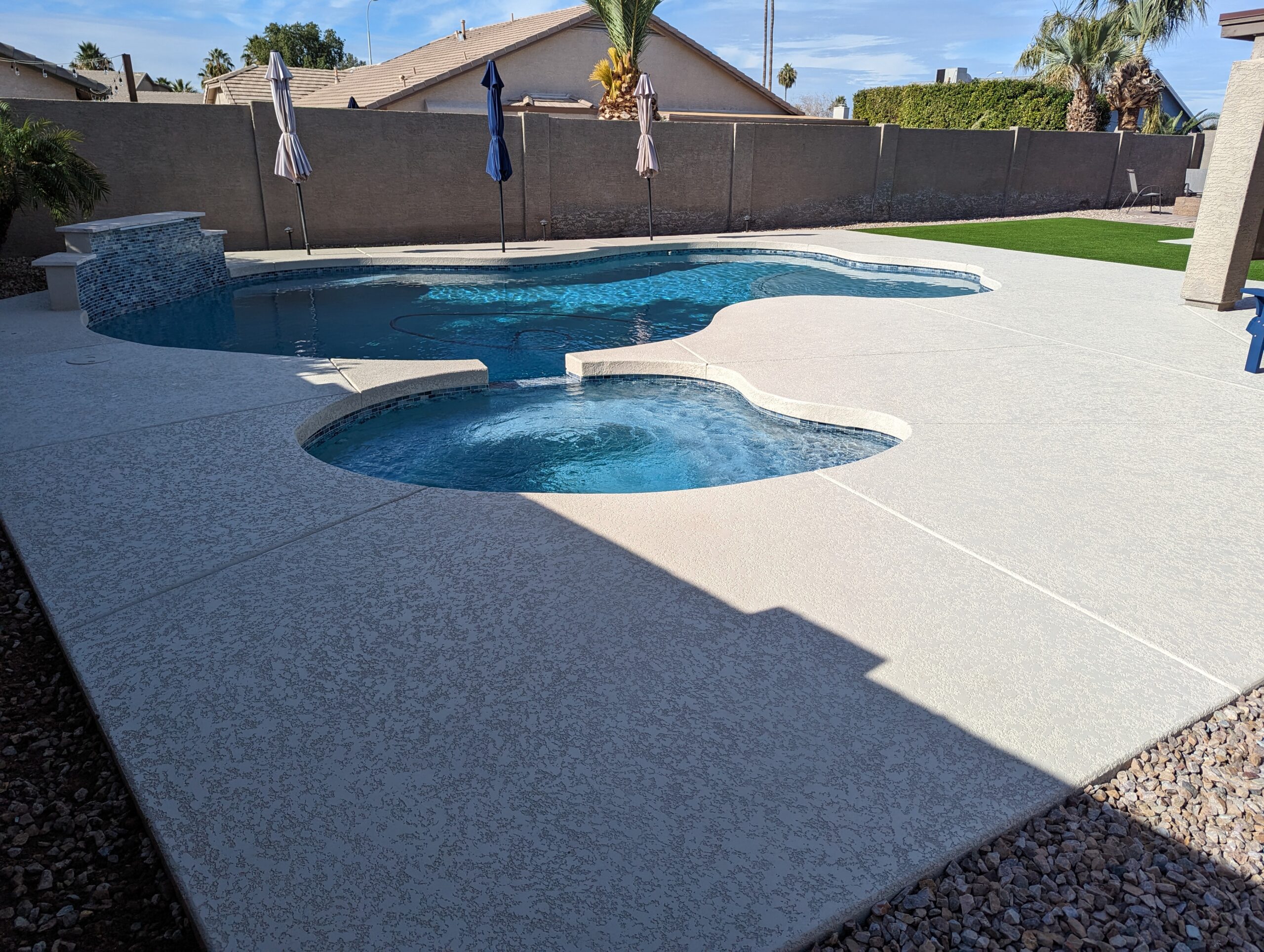 Pool area featuring a sparkling blue swimming pool and spa, surrounded by a textured concrete deck, with patio umbrellas and landscaping, showcasing a well-maintained residential outdoor space in Gilbert, AZ.