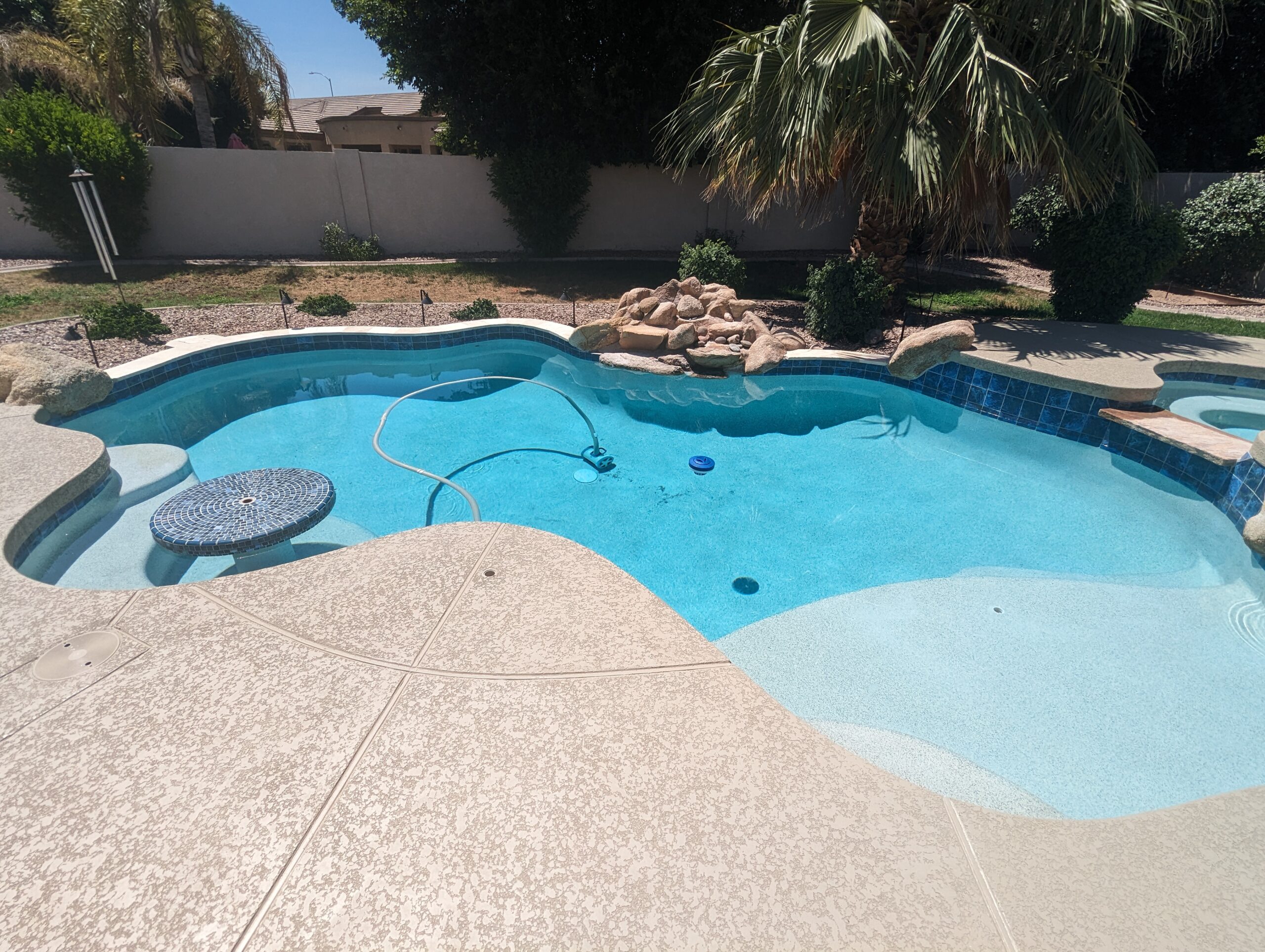 Sparkling blue swimming pool with a decorative stone feature, surrounded by a clean deck and lush greenery, showcasing Pelican Pools' expert maintenance and service quality.