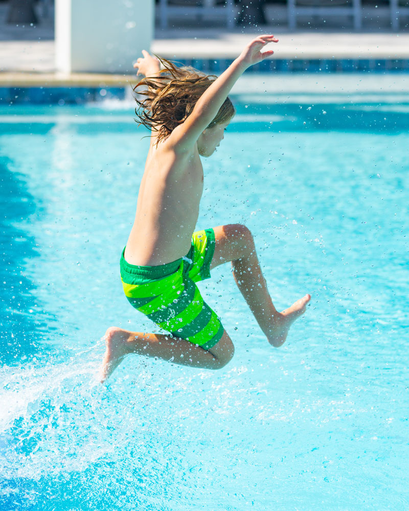 Child in green striped swim trunks jumping into a pool, creating splashes of water, representing fun and enjoyment in a residential pool setting.