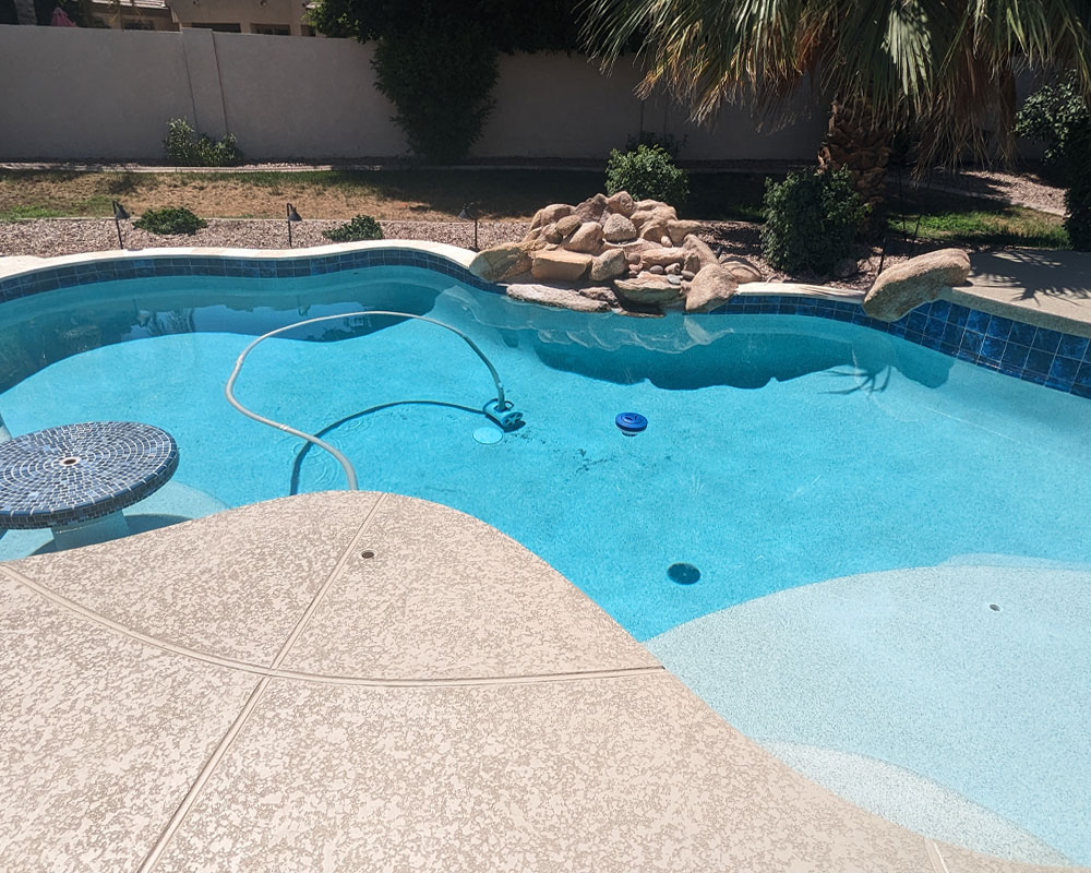 Swimming pool with clear blue water, featuring a mosaic-topped table, pool cleaning equipment, and landscaped surroundings, illustrating Pelican Pools' weekly maintenance services for homeowners in Arizona.