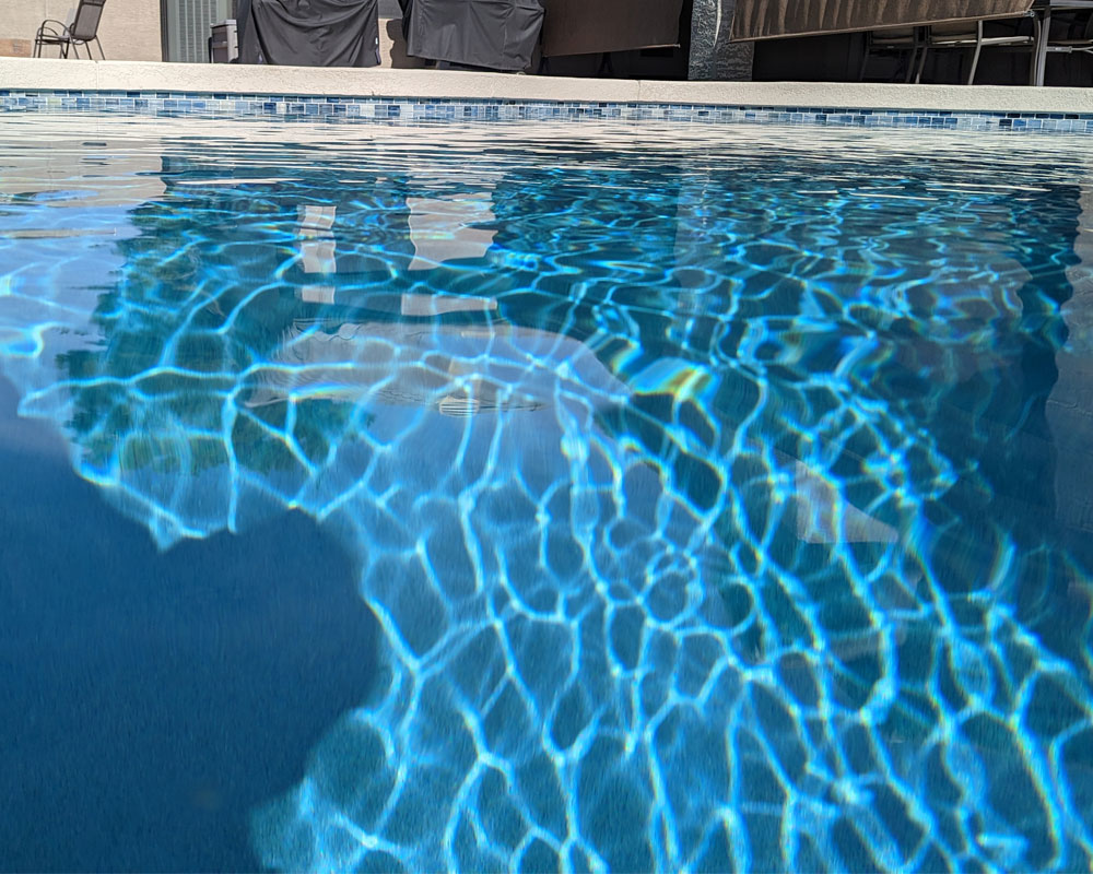 Clear swimming pool water reflecting sunlight, showcasing rippling surface patterns, with poolside furniture in the background, relevant to Pelican Pools' services in pool maintenance and diagnostics.