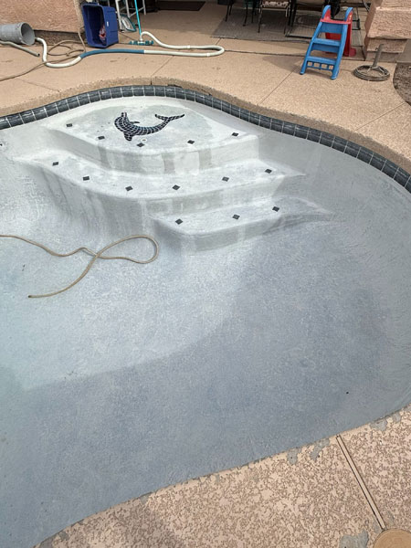 Pool with light-colored surface, showing steps and decorative tile, indicating recent cleaning process; surrounding area includes pool equipment and patio furniture, reflecting Pelican Pools' green to clean service.