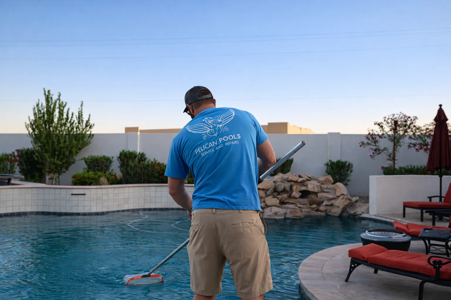 Pool service technician from Pelican Pools cleaning a residential pool in Arizona, surrounded by landscaped garden and patio furniture, emphasizing pool maintenance and care.